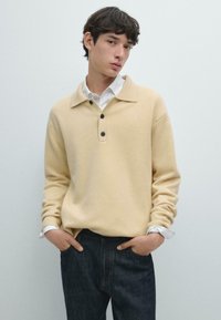 Pullover a maglia giallo chiaro con colletto e tre bottoni scuri. Maniche lunghe con risvolti su una camicia bianca, abbinato a jeans scuri.