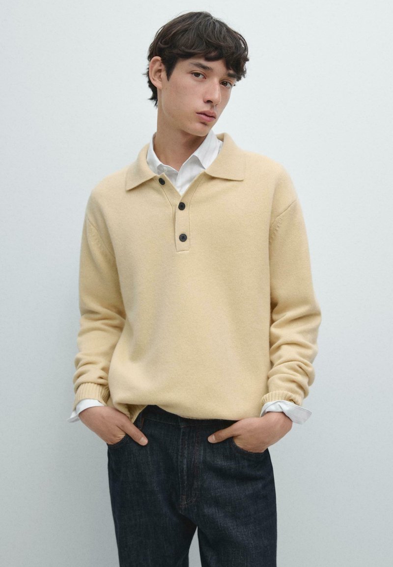 Pullover a maglia giallo chiaro con colletto e tre bottoni scuri. Maniche lunghe con risvolti su una camicia bianca, abbinato a jeans scuri.
