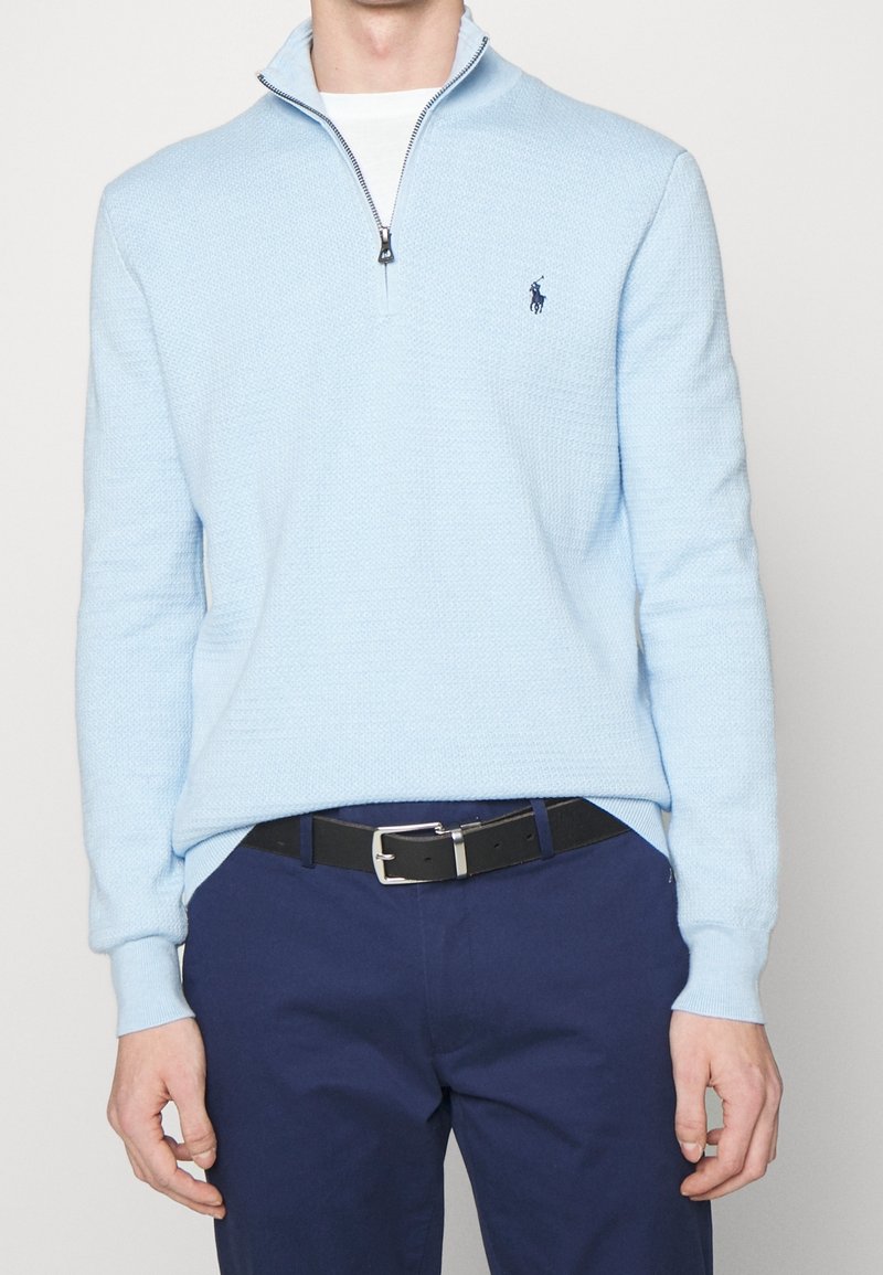 Homme portant un pull bleu clair à fermeture quart de zip, une chemise blanche en dessous, un pantalon bleu marine et une ceinture noire avec une boucle argentée.