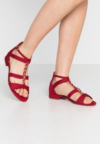 Jalat yang mengenakan sandal merah bertali dengan detail rantai emas, berdiri di atas lantai putih yang halus dengan satu kaki sedikit terangkat.