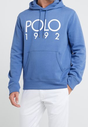 Mann trägt blauen Hoodie mit dem Schriftzug „POLO 1992“ und weiße Hose, Hände sichtbar, vor einfachem hellem Hintergrund.