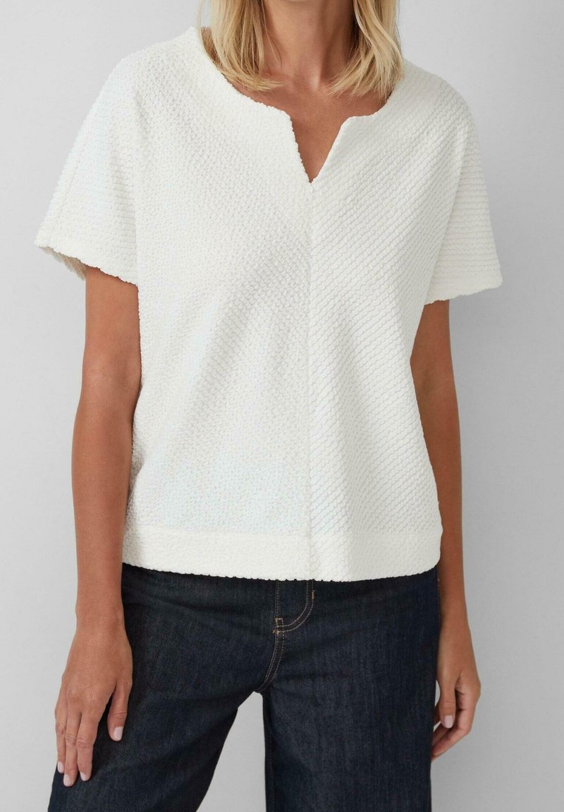 Femme portant un haut blanc texturé à manches courtes avec un col en V, associé à un jean en denim bleu foncé.