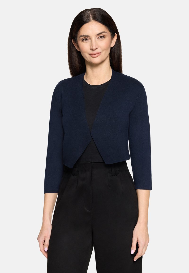Frau mit glattem dunklem Haar trägt eine marineblaue kurze Strickjacke über einem schwarzen Top und schwarzen Hosen mit hoher Taille, stehend vor weißem Hintergrund.