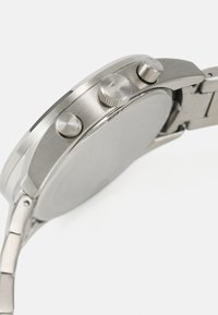 Ceas din oțel inoxidabil argintiu, cu carcasă rotundă și două butoane de presiune, având o coronă texturată și un design cu brățară din lanț.