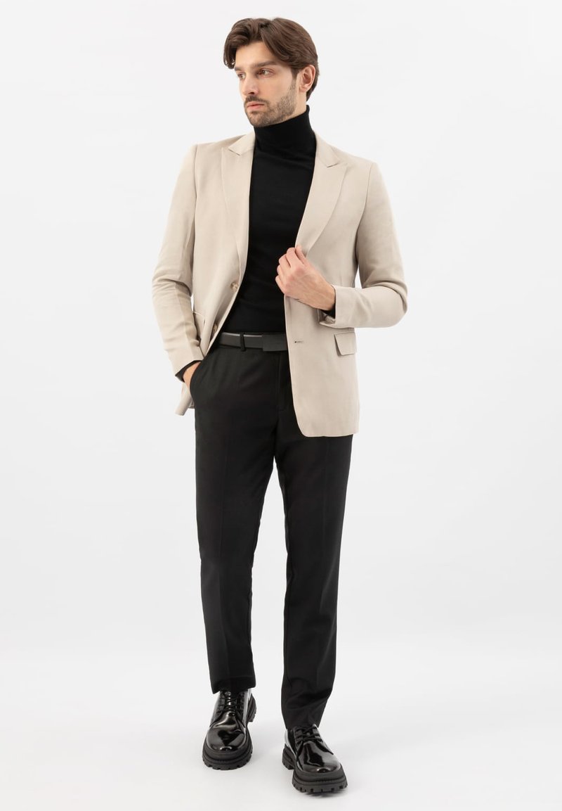 Mężczyzna w beżowej marynarce, czarnym golfie, czarnych spodniach i czarnych błyszczących skórzanych butach, stojący z jedną ręką w kieszeni.