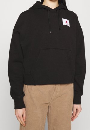 Personne portant un sweat à capuche noir avec un patch logo blanc et rouge sur la poitrine et un pantalon en velours côtelé beige sur un fond uni.