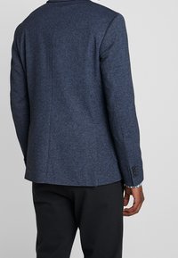 Tmavě modrý texturovaný blazer s přiléhavým střihem, se zadním ventilem a černými knoflíkovými akcenty na manžetách. Nosený s tmavými kalhotami.