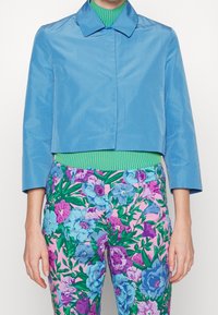 Giacca blu corta con colletto classico, abbinata a un dolcevita a righe verdi e a pantaloni con motivo floreale nei toni del rosa, blu e verde.