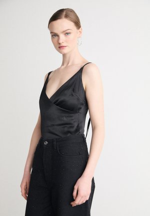Camisole nera in satin con design a V, caratterizzata da spalline sottili e un logo discreto. Abbinata a pantaloni neri decorati con piccoli chiodini.