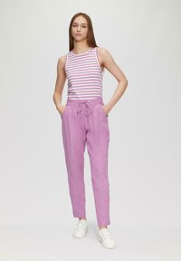 Paarse linnen broek met elastische tailleband en koord. Gestreept mouwloos topje in lichtroze en wit. Model staat met de handen in de zakken.