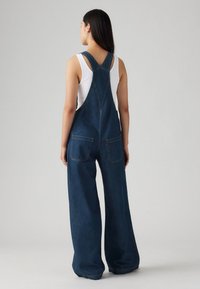 Denim jumpsuit med vide ben, justerbare skulderstropper og to baklommer. Fargen er mørkeblå med synlige sømdetaljer.