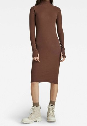 Femme portant une robe midi ajustée marron côtelée à manches longues avec des trous pour les pouces, associée à des bottines beiges à lacets et des chaussettes marron.