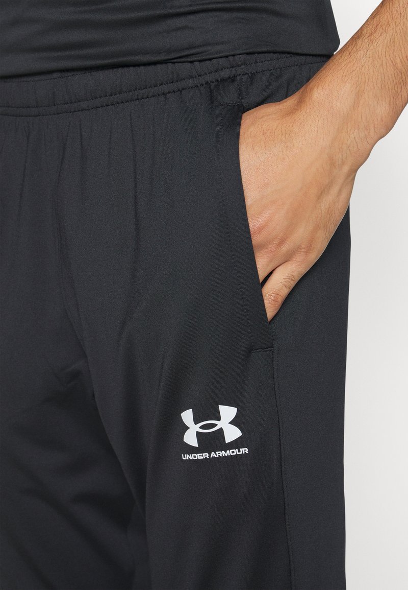 Czarne spodnie sportowe wykonane z gładkiego, lekkiego materiału, z elastycznym paskiem w talii i kieszeniami bocznymi. Posiadają białe logo Under Armour.