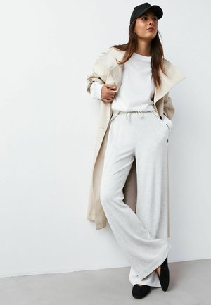 Beiger Trenchcoat, weißes langärmliges Hemd, hellgraue weit geschnittene Hose mit Kordelzug und schwarze Slip-on-Schuhe vor weißem Hintergrund.