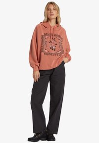 Felpa color ruggine con grafica floreale e testo del marchio. Il modello la indossa con pantaloni a gamba larga neri e stivaloni neri spessi.