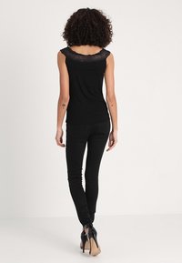 Top negro sin mangas con ribete de encaje en el escote, combinado con jeans ajustados negros y zapatos negros de tacón alto. Cabello rizado visible.