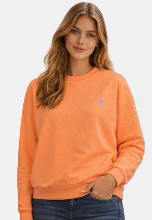 Woman with long blonde hair wearing an orange sweatshirt and blue jeans, standing with one hand in her pocket against a plain background.