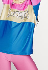 Personne portant un maillot de sport coloré avec le texte "HEY SPORTS" et des leggings roses, levant les bras, avec des cheveux tressés visibles d'un côté.