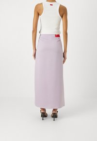 Lila maxi rok met een gladde textuur, een aangesloten taille en een rode labeldetail. Draagt een witte tanktop en zwarte hoge hakken.