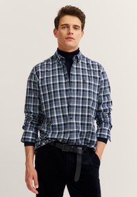Camicia a quadri da uomo con motivo a scacchi blu navy e bianco, maniche lunghe e colletto arrotondato; indossata sopra un dolcevita nero.
