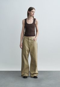 Kvinne stående med hånden i lommen, iført en brun ermeløs topp og vidbente beige bukser med et subtilt prikkemønster på en ensfarget grå bakgrunn.