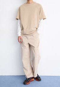 Camiseta de manga corta beige superpuesta sobre una camisa de manga larga blanca, combinada con pantalones cargo beige claros y zapatillas negras con detalles en rojo.
