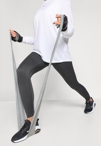 Čierne športové legíny s rebrovanými detailmi, v kombinácii s voľným bielym tričkom s dlhým rukávom a čiernymi športovými topánkami. V ruke drží odporovú gumu.