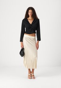 Svart crop-top med lange ermer og vridd front, kombinert med en plisserte, metallisk gull midi-skjørt, og svart håndveske. Hvite sandaler med stropper.