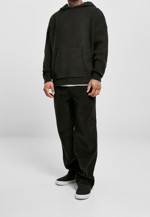 Zwarte geribbelde hoodie met een voorzak, gecombineerd met zwarte corduroy broek en zwarte schoenen met witte zolen. Materiaal lijkt zacht en textuur te hebben.