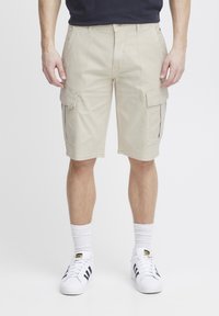 Beige cargo sort, több oldalsó zsebel, övhurkok és egyenes szabás. Fehér zoknikkal és fekete csíkos sportcipővel viselve.