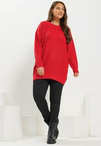 Femme portant un gros pull rouge, un pantalon noir et des bottes noires, se tenant avec une jambe croisée devant un fond blanc.