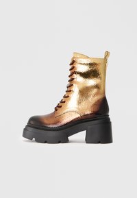 Botte de combat à lacets avec finition en or métallique et marron, surface texturée, talon noir épais et tirette.