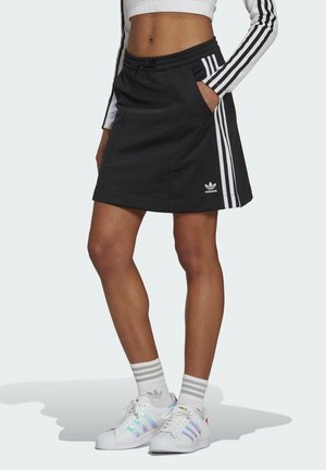 Kobieta nosząca czarną spódnicę Adidas z białymi paskami i logo, białe skarpetki w paski oraz białe buty sportowe Adidas z paskami w kolorach tęczy.