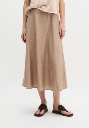 Kobieta ubrana w beżową midi spódnicę i bezrękawnik z dzianiny, w połączeniu z czarnymi sandałami z odkrytymi palcami, stojąca na jednolitym tle.