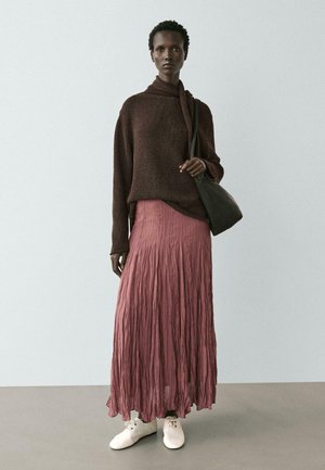 Kobieta ubrana w brązowy dziergany sweter, długą pogniecioną fioletowawą spódnicę, białe sznurowane buty i niosąca czarną torebkę na ramię na tle gładkiej ściany.