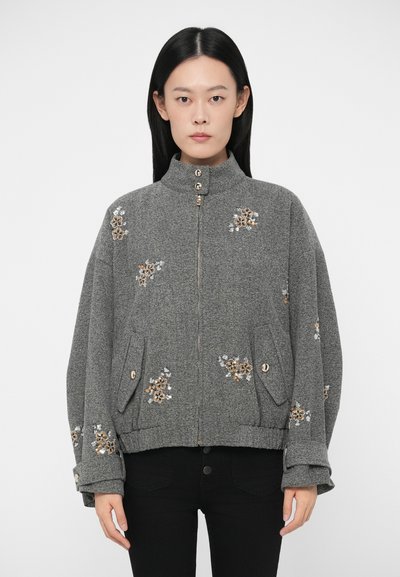 Femme portant une veste grise zippée avec des poignets boutonnés et des embellissements en sequins floraux, associée à un pantalon noir, se tenant devant un fond uni.
