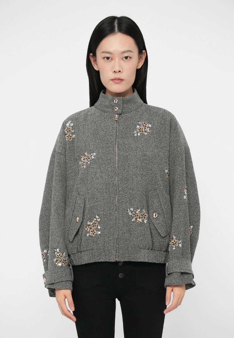 Sieviete, ģērbta pelēkā ar rāvējslēdzēju un pogām rotātā jakā ar ziedu aplikācijām, valkā melnas bikses un stāv pret vienkāršu fona.