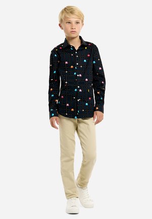Camisa de manga larga de algodón, con un fondo negro y patrones coloridos de Pac-Man y fantasmas. Combinada con pantalones beige y zapatos blancos.
