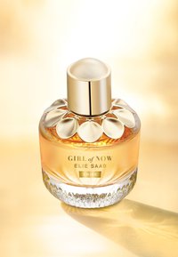 Okrągła szklana butelka perfum z złotą kwiatową zakrętką, zawierająca bursztynowy płyn; przezroczysta podstawa z teksturowanymi detalami; etykieta z napisem "GIRL of NOW ELIE SAAB SHINE."