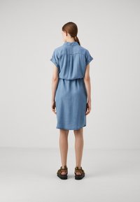 Ljusblå denimklänning med korta ärmar, lätt rynkad midja och rak skärning, tillsammans med bruna sandaler.
