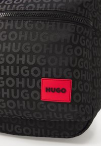 Fekete hátizsák, amelyen sötétebb árnyalatban ismétlődő "HUGO" minta található. A táskán feltűnő piros logó folt van fekete szöveggel, valamint cipzáras zseb.