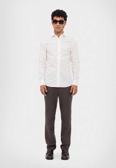 Witte, lange mouwen knopshirt, smalle manchetten, gecombineerd met donker gestreepte broeken en zwarte dresschoenen. Het model draagt donkere zonnebrillen.