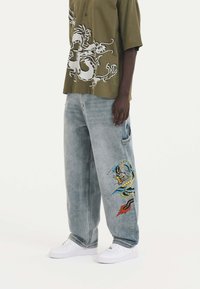 Ljusblå denimbyxor med en lös passform, med en färgglad drakebroderi på vänster ben. Matchas med en olivgrön skjorta.