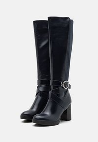Botas de charol azul marino hasta la rodilla, con un tacón ancho y una correa ajustable con un detalle de hebilla metálica circular.