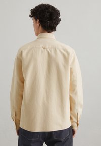 Light yellow long-sleeve shirt with a textured fabric, buttoned cuffs, and back yoke detail, paired with dark denim jeans.