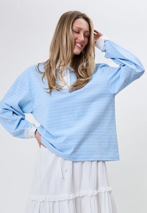 Smilende kvinde med langt blondt hår iført en lyseblå stribet sweater over en hvid flæset nederdel, poserer med den ene hånd på hoften og lukkede øjne.