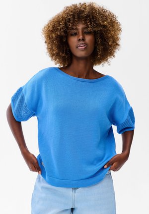 Kobieta o kręconych włosach ubrana w jasnoniebieski dzianinowy top z siateczkowymi rękawami i jasnoniebieskie dżinsy, stoi z rękami na biodrach.