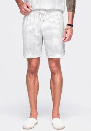 Mand iført hvide shorts med tynde striber og snøre samt hvide slip-on sko, står foran en ensfarvet baggrund.