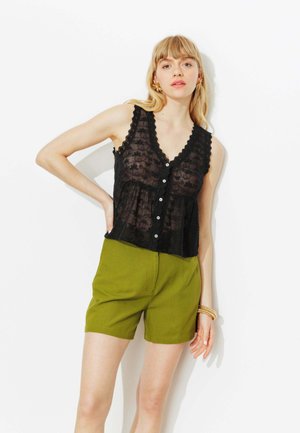 Femme aux cheveux blonds portant un haut sans manches transparent noir à boutons et un short vert olive, debout devant un fond blanc.