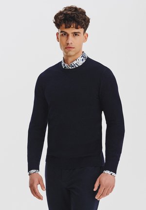 Granatowy sweter z okrągłym dekoltem o fakturowanym splocie, nałożony na koszulę z wzorzystym kołnierzykiem. Całość ma elegancki i dopasowany fason.